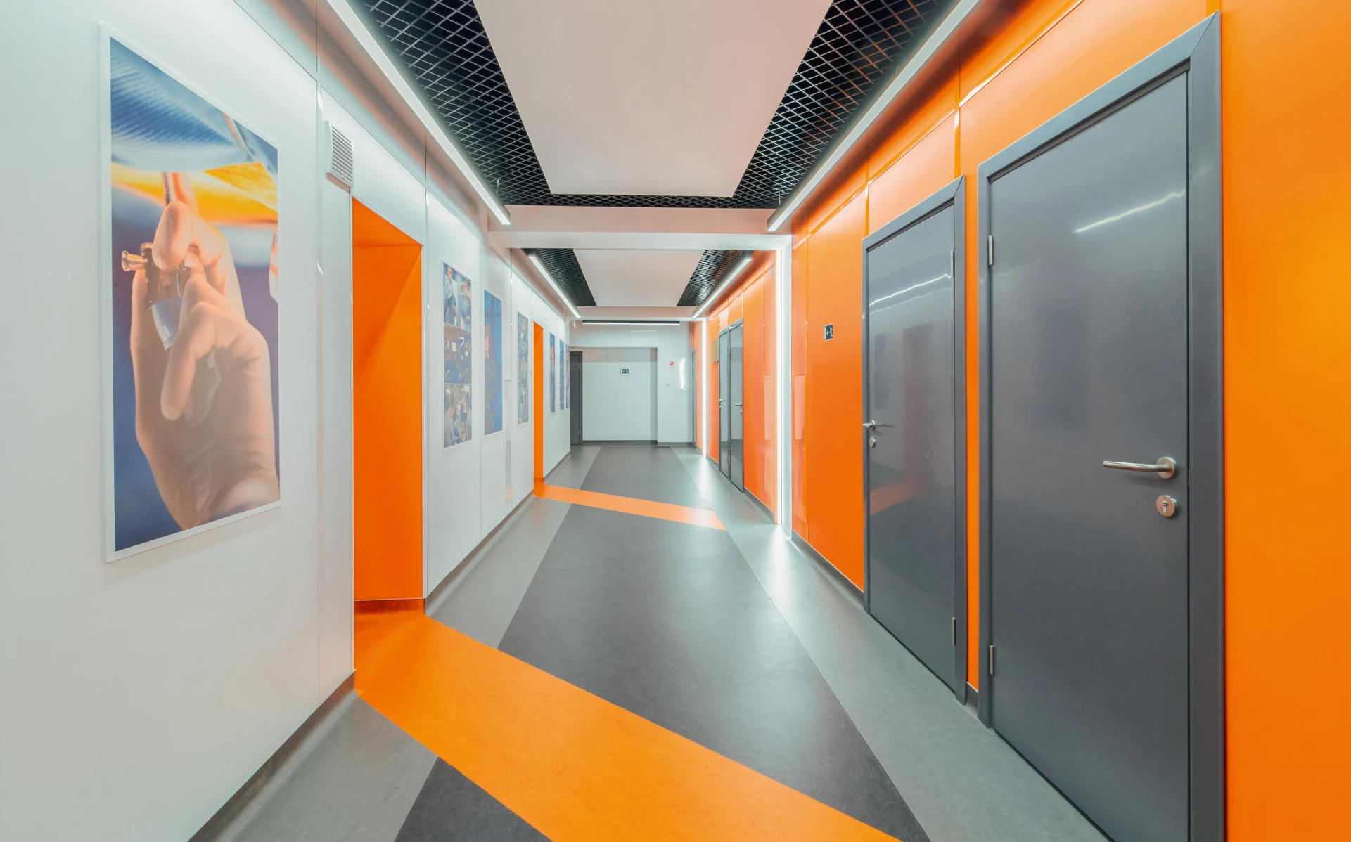 St George training center corridor: light walls, gray doors, orange accents on walls and floors, and linear lighting along a black open-grid ceiling